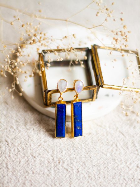 Lapis and moonstone earrings with gold accents on a white coaster with dried flowers.