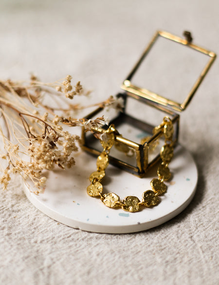 Gold bracelet on a white coaster with dried flowers and a small glass jewellery box and dried flowers in the background.