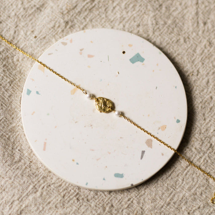 Gold bracelet with a gold charm and pearl beads on a round, speckled coaster 