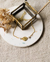 Gold bracelet with a gold charm and pearl beads on a speckled white coaster with dried flowers.