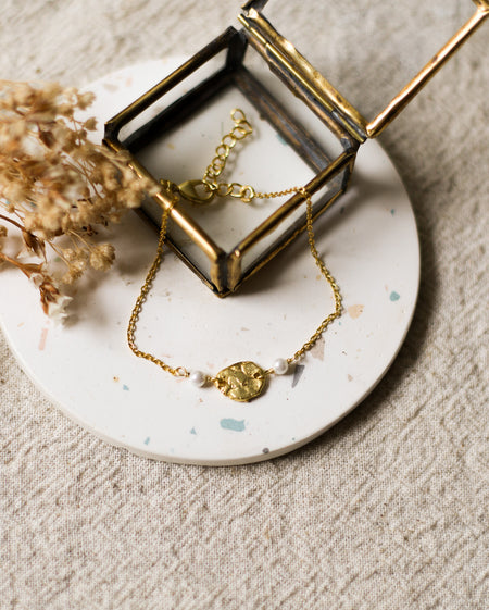 Gold bracelet with a gold charm and pearl beads on a speckled white coaster with dried flowers.