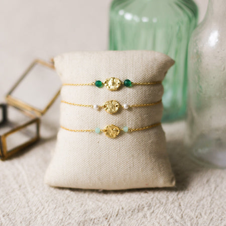 Three gold bracelets with gemstone beads on a beige pillow, with two glass bottles in the background.