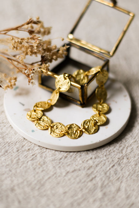Gold bracelet on a white speckled coaster with a glass jewellery box and dried flowers in the background