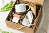 Kraft gift box containing a cream mug, chocolate bar, forged brass spoon and mini bouquet of flowers, with tulips on a beige linen background