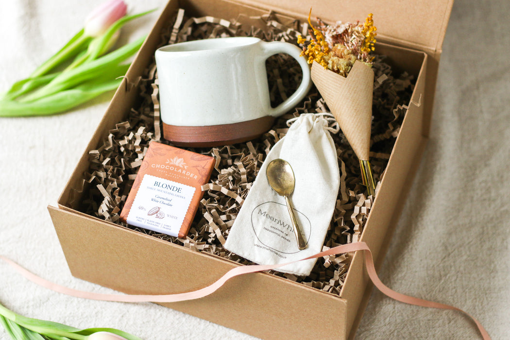 Kraft gift box containing a cream mug, chocolate bar, forged brass spoon and mini bouquet of flowers, with tulips on a beige linen background