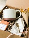 Kraft gift box containing a cream mug, chocolate bar, forged brass spoon and mini bouquet of flowers, on a beige linen background