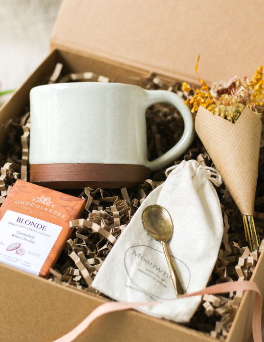 Kraft gift box containing a cream mug, chocolate bar, forged brass spoon and mini bouquet of flowers, on a beige linen background