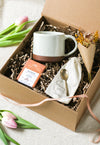 Kraft gift box containing a cream mug, chocolate bar, forged brass spoon and mini bouquet of flowers, with tulips on a beige linen background
