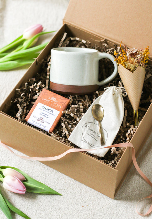 Kraft gift box containing a cream mug, chocolate bar, forged brass spoon and mini bouquet of flowers, with tulips on a beige linen background