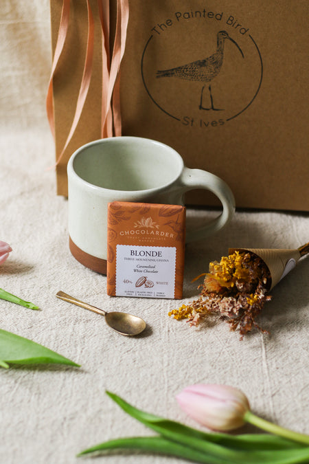 Kraft gift box with a cream mug, chocolate bar, forged brass spoon and mini bouquet of flowers, with tulips on a beige linen background