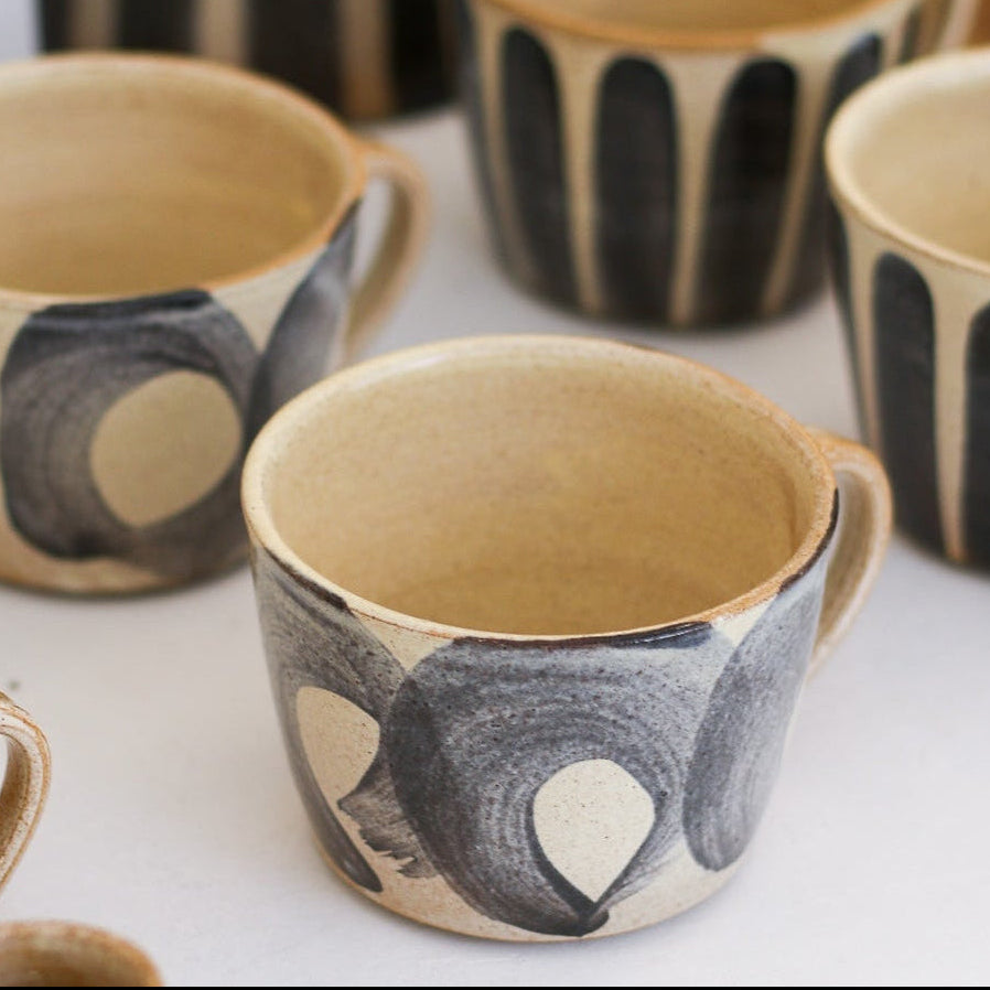 Set of beige stoneware cups with hand painted blue stripes and patterns on a white surface with a blurred background