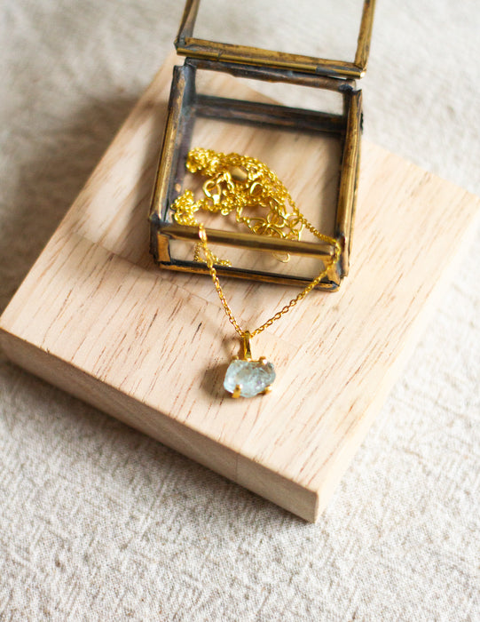Gold necklace with an aqua pendant in an open brass jewellery box on a wooden and linen surface.
