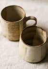 Two beige speckled ceramic mugs with a textured surface on a light fabric background