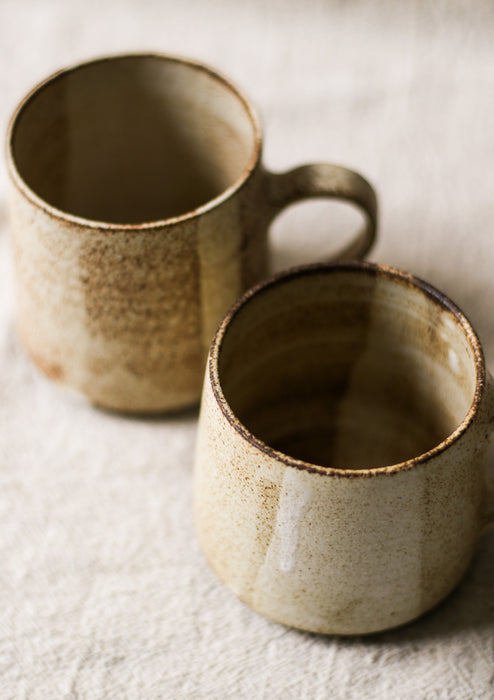 Two beige speckled ceramic mugs with a textured surface on a light fabric background