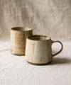 Two beige speckled ceramic mugs on a textured surface with a neutral background