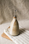 Ceramic oil bottle with a cork and metal pourer, on a beige striped tea towel and wooden board, with a beige linen background