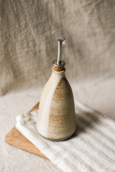 Ceramic oil bottle with a cork and metal pourer, on a beige striped tea towel and wooden board, with a beige linen background