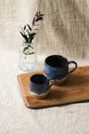 Two blue ceramic mugs on a wooden tray with a glass jar containing greenery in the background.