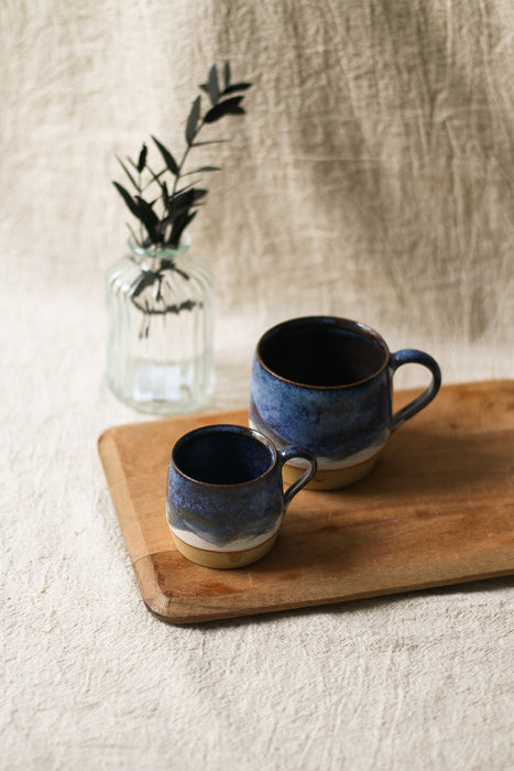 Two blue ceramic mugs on a wooden tray with a glass jar containing greenery in the background.
