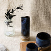 Two blue ceramic mugs and a blue vase on a wooden surface with a textured beige wall background.