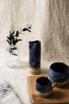 Two blue ceramic mugs and a blue vase on a wooden surface with a textured beige wall background.