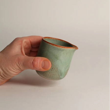 Hand holding a small green ceramic jug against a plain grey background