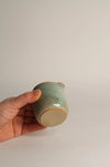 Hand holding a green ceramic jug against a plain grey background