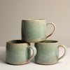 Three green ceramic mugs, stacked, on a light grey background