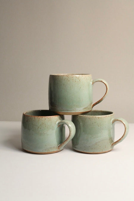 Three green ceramic mugs, stacked, on a light grey background