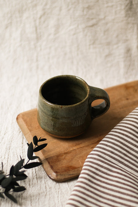 Green ceramic mug on a wooden tray with a striped cloth and leaves in the background