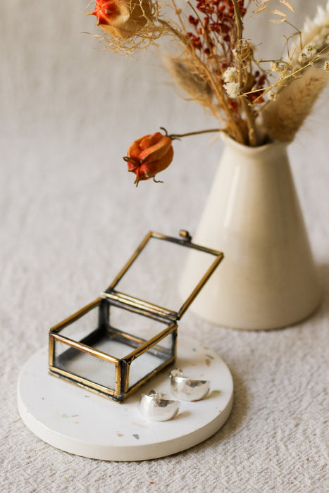 A small open jewellery box with a brass frame and clear glass lid sits on a round white coaster beside two silver crescent shaped earrings. A cream vase with dried flowers is blurred in the background on a light fabric surface.