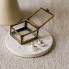 A pair of silver crescent-shaped earrings displayed on a round white terrazzo coaster next to a small brass framed glass jewellery box, set on a textured beige linen surface.