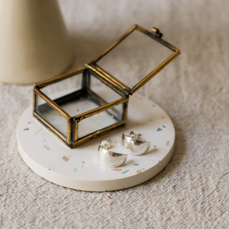 A pair of silver crescent-shaped earrings displayed on a round white terrazzo coaster next to a small brass framed glass jewellery box, set on a textured beige linen surface.