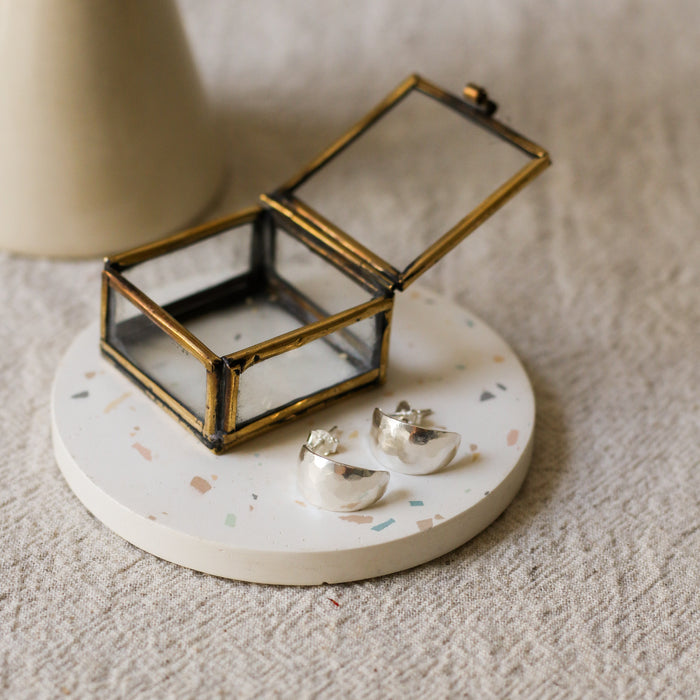 A pair of silver crescent-shaped earrings displayed on a round white terrazzo coaster next to a small brass framed glass jewellery box, set on a textured beige linen surface.