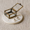 A pair of silver crescent-shaped earrings displayed on a round white terrazzo coaster next to a small brass framed glass jewellery box, set on a textured beige linen surface.