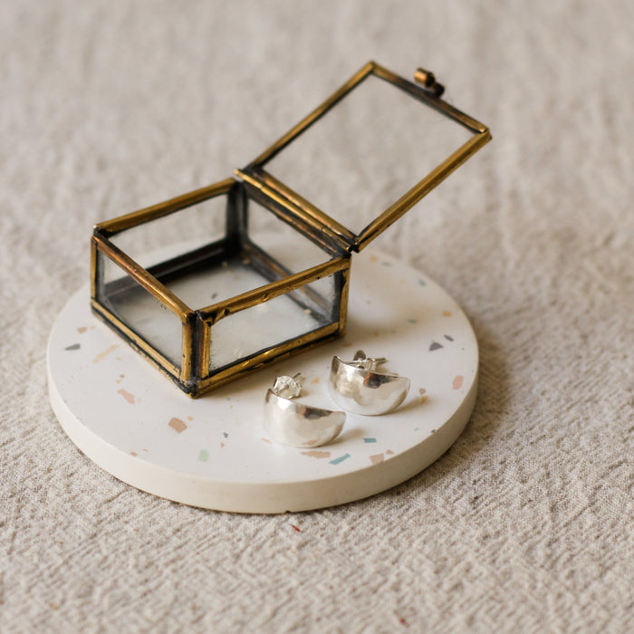 A pair of silver crescent-shaped earrings displayed on a round white terrazzo coaster next to a small brass framed glass jewellery box, set on a textured beige linen surface.