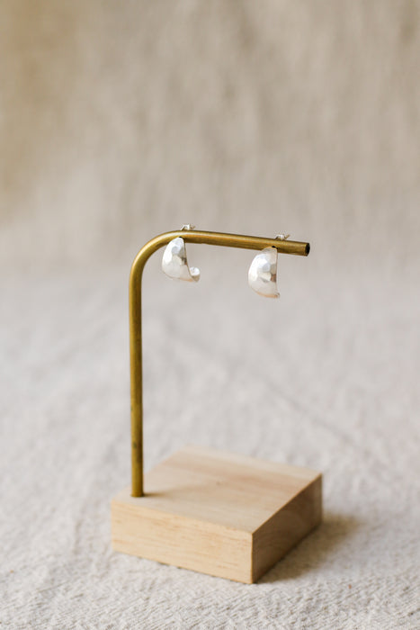 A pair of silver crescent-shaped stud earrings displayed on a brass stand with a light wood base against a soft beige linen background.