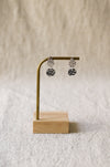 A pair of silver teardrop dangle earrings with textured, faceted drop pendants, displayed on a small brass stand with a wooden base against a soft beige textured background.