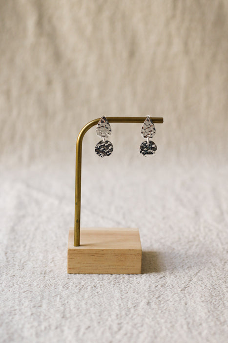 A pair of silver teardrop dangle earrings with textured, faceted drop pendants, displayed on a small brass stand with a wooden base against a soft beige textured background.