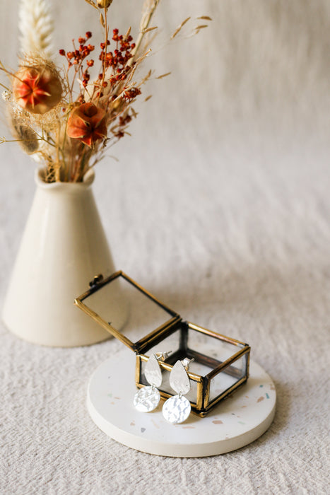 Silver teardrop-shaped drop earrings with hammered disc pendants, displayed in a small brass jewellery box on a round white coaster, with a blurred vase of dried flowers in the background.