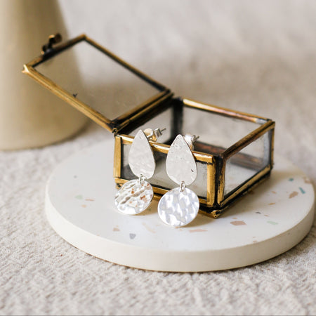Silver teardrop-shaped drop earrings with hammered disc pendants, displayed in a small brass jewellery box on a round white coaster, with a blurred vase in the background.