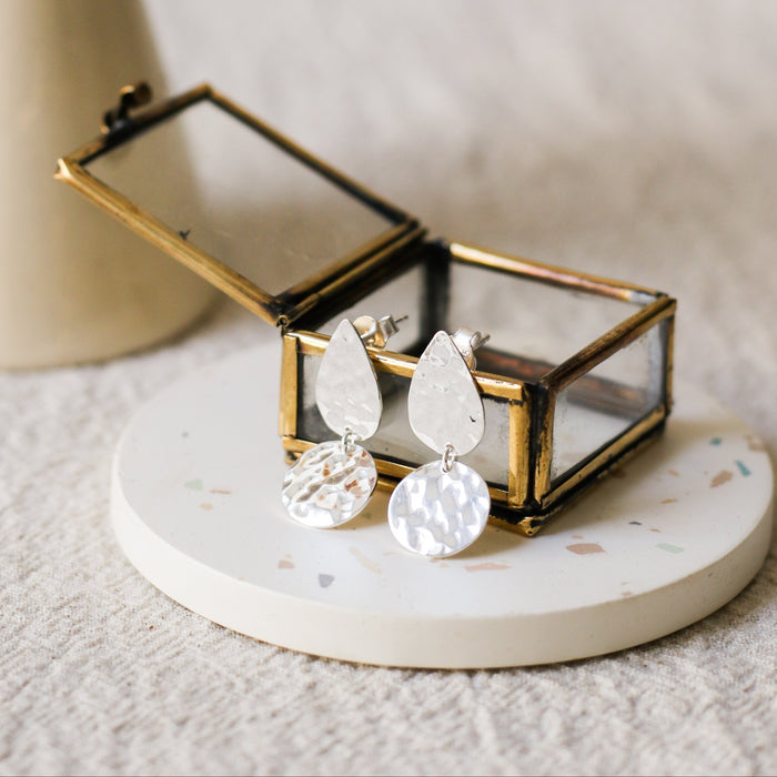 Silver teardrop-shaped drop earrings with hammered disc pendants, displayed in a small brass jewellery box on a round white coaster, with a blurred vase in the background.