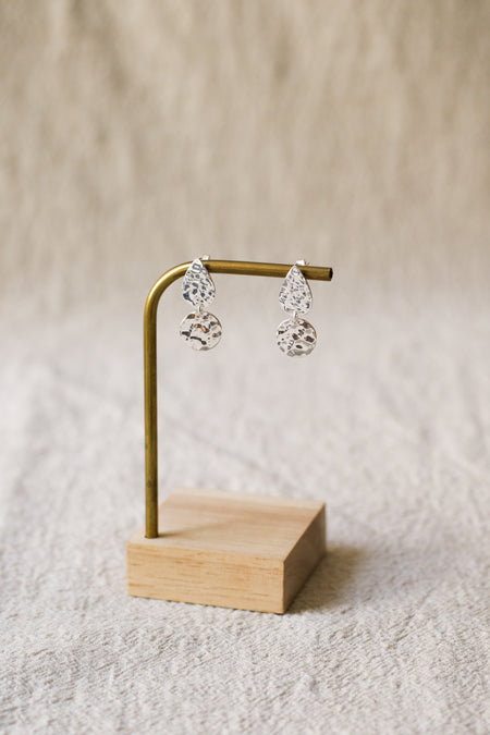 A pair of silver teardrop dangle earrings with textured, faceted drop pendants, displayed on a small brass stand with a wooden base against a soft beige textured background.