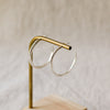 A pair of silver hoop earrings displayed on a brass stand with a light wood base against a soft beige textured background.