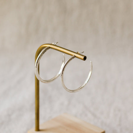 A pair of silver hoop earrings displayed on a brass stand with a light wood base against a soft beige textured background.