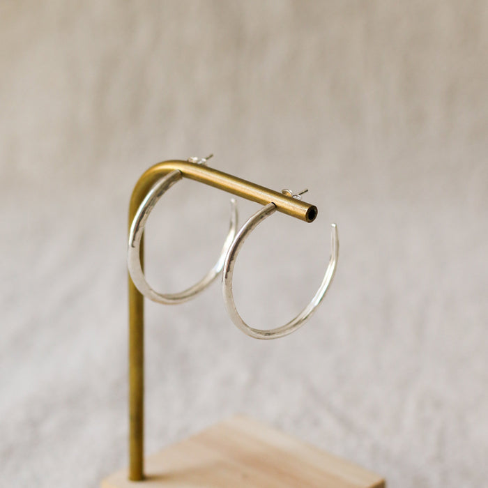 A pair of silver hoop earrings displayed on a brass stand with a light wood base against a soft beige textured background.