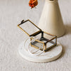 A small open jewellery box with a brass frame and clear glass lid sits on a round white coaster. Two silver hoop earrings are draped over the front.