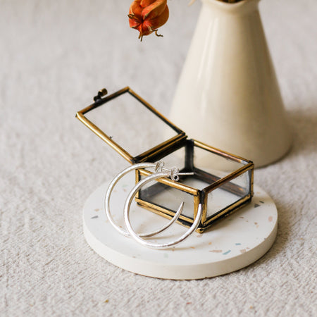 A small open jewellery box with a brass frame and clear glass lid sits on a round white coaster. Two silver hoop earrings are draped over the front.