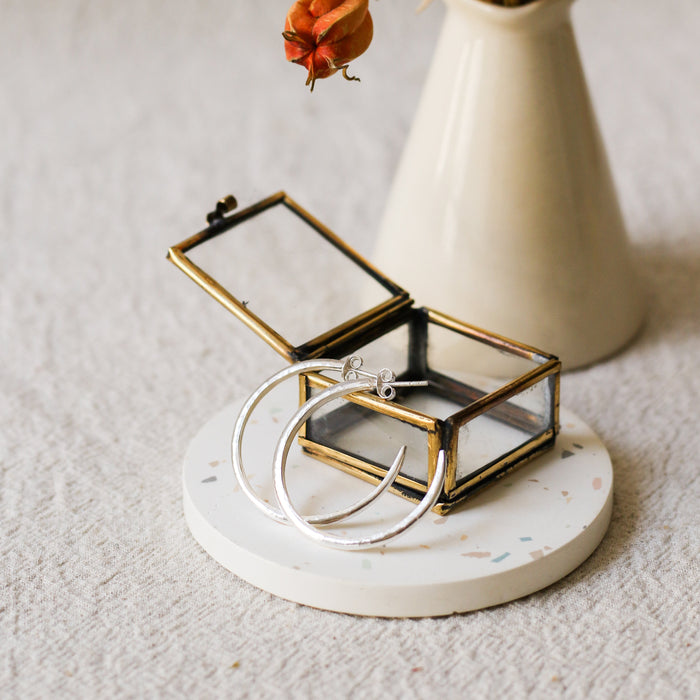 A small open jewellery box with a brass frame and clear glass lid sits on a round white coaster. Two silver hoop earrings are draped over the front.