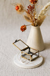 A small open jewellery box with a brass frame, holding two silver hoop earrings, styled on a round white coaster with dried flowers in a cream vase behind it.